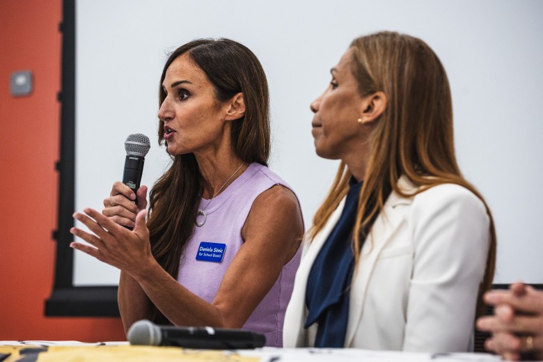 A photograph by Dave Decker showing Hillsborough County School Board candidate Daniela Simic speaking at the C. Blythe Andrews Jr. Public Library in Tampa, Florida, on July 9, 2026. Captured in a profile shot from the side, Simic holds a microphone in her right hand while gesturing with her left hand. She wears a sleeveless lilac top, a blue campaign button reading "Daniela Simic for School Board" on her chest, a gold necklace, and a circular pendant. Seated to her right is another panelist wearing a white blazer over a dark navy top, listening as Simic addresses the room.