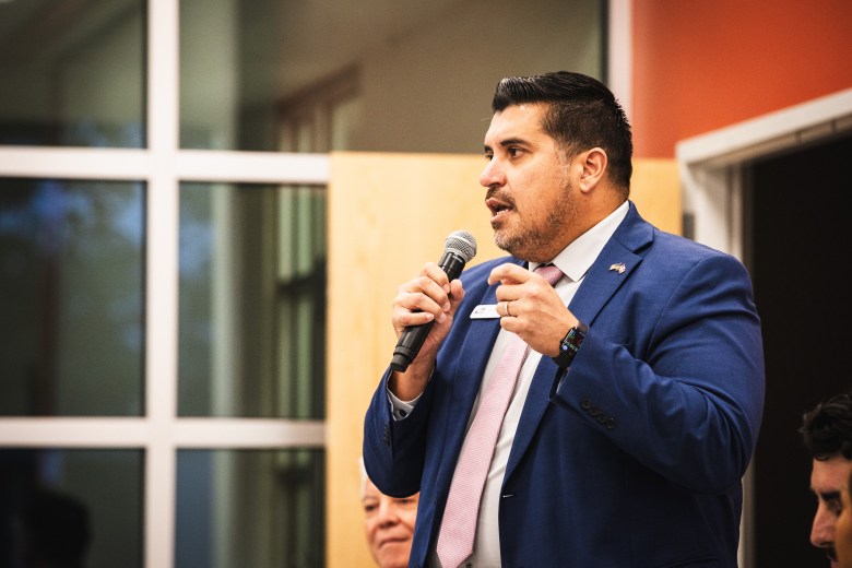 A photograph by Dave Decker showing Luis Salazar speaking at the C. Blythe Andrews Jr. Public Library in Tampa, Florida, on July 9, 2026. Captured in a medium three-quarter shot, Salazar holds a microphone in his right hand while gesturing with his left hand as he addresses the audience. He wears a tailored blue suit with a lapel pin, a white collared shirt, and a pink tie against a background featuring large framed glass windows.