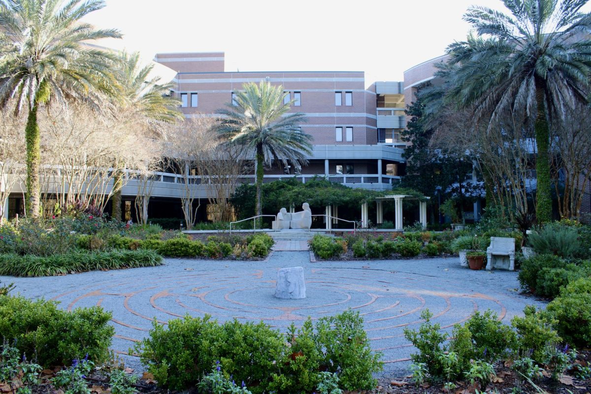 A courtyard featuring a brick-lined circular labyrinth set into a light gravel surface in the foreground, with a small stone block at its center. Low green shrubs and various plants border the labyrinth. In the middle ground, steps lead up to a white, abstract sculpture of two figures sitting together, framed by palm trees and bare branches. A large, multi-story brick building with horizontal gray bands and windows serves as the backdrop under a clear sky.