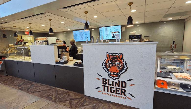 A photograph by Ray Roa showing the ordering counter and interior coffee shop setup of Blind Tiger Coffee Roasters inside the Rivergate Tower in downtown Tampa, Florida, on August 11, 2026. Captured in a wide angle shot, the foreground prominently features a white ordering counter displaying a large tiger head logo alongside black lettering reading "BLIND TIGER COFFEE ROASTERS." Behind the counter, digital menu boards hang beneath recessed drop-ceiling lights, flanking a glass pastry display case and retail merchandise on display under bright interior lighting.