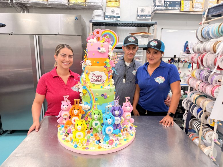 A photograph showing bakery owner Kristina Lavallee and two staff members posing behind a tall, multi-tiered Care Bears themed custom birthday cake created for Cardi B inside The Cake Girl bakery kitchen. Positioned center frame around a stainless steel prep table, Lavallee stands on the left in a bright pink collared polo shirt, while two team members in blue polo shirts and black logo caps stand to the right. The colorful multi-tiered pastel cake features fondant Care Bear figurines around its base, floral and rainbow decorations, a pink number "6" topper, and a white plaque reading "Happy Birthday." Shelves filled with cake boxes, ribbon spools, and commercial kitchen equipment fill the background under bright indoor lighting.