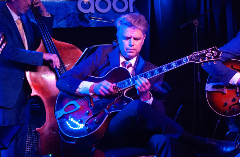 A photograph of jazz guitarist Carl Amundson performing live under vibrant blue stage lighting. Seated centrally in a dark suit jacket, white collared shirt, red patterned tie, and dark trousers with his legs crossed, Amundson focuses intently while playing a sunburst Guild hollowbody electric guitar. On the left behind him, a upright bass player in a dark suit with gold sleeve stripes plays a double bass, while a silver sign reading "door" is visible in the background.