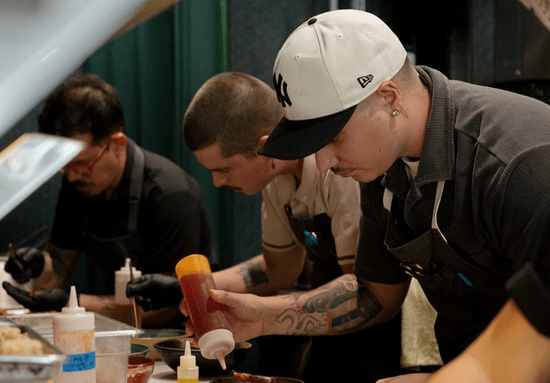 A photograph by Kayla Surico showing Chef Yariel Berrios and kitchen staff plating dishes inside Coro Restaurant in Orlando, Florida, on April 14, 2026. Captured in a medium close-up profile shot, Chef Berrios stands on the right wearing a white New York Yankees baseball cap with a black visor, a dark grey shirt, black apron, and black gloves, focusing closely as he dispenses red sauce onto a dish from a plastic squeeze bottle. To his left, two kitchen team members in black aprons and gloves work side-by-side along the prep line under warm, directional kitchen lighting.