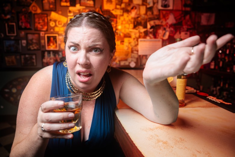 A promotional character portrait by James Zambon featuring actor Colleen Cherry as Penelope for Jobsite Theater's upcoming production of "Penelope." Captured in a medium close-up shot leaning over a wooden bar top inside a dimly lit venue, Cherry wears a dark teal sleeveless dress, a layered gold chain necklace, and braided hair pinned around her head. Looking directly into the camera lens with a skeptical facial expression, she holds a rocks glass filled with amber liquor in her right hand while gesturing outwards with her open left hand. In the background, framed photos, posters, and warm orange accent lighting create an intimate barroom atmosphere, although the photo is shot inside The Hub dive bar in downtown Tampa.