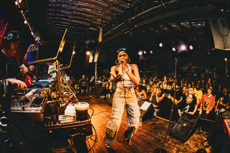 A wide-angle concert photograph showing hip-hop artist Dynasty performing live on stage during a concert at Crowbar in Ybor City, Florida, on July 31, 2026. Captured in a full-length shot from the left side of the stage, Dynasty stands centered under warm stage lights holding a microphone to her mouth, wearing a white crop top, silver hoop earrings, and wide-leg light blue denim cargo pants. To the left, DJ Sandman, widely considered the godfather of Tampa hip-hop, operates mixing equipment and a laptop behind a elevated table setup, while a crowded audience stands in front of the stage with hands raised in the background under the dark, exposed-beam venue ceiling.