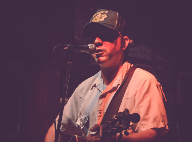 A concert photograph by Nicole Kibert showing singer-songwriter David Dondero performing live on stage at New World Brewery in Ybor City, Florida, on November 23, 2013. Captured in a medium close-up shot under warm red stage lighting, Dondero stands singing into a vocal microphone while playing an acoustic guitar. He wears a dark baseball cap with a gold emblem over dark sunglasses, paired with a short-sleeved button-up shirt featuring light blue and white color-blocked panels against a dark, moody background.