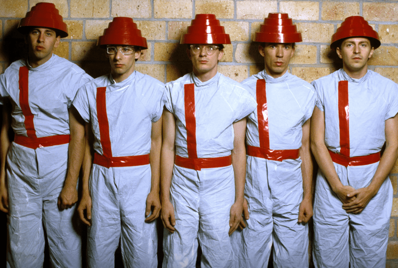 Promotional photo for Chris Smith's Devo documentary showing the five members of the new wave band Devo posing in front of a tan brick wall. From left to right, the members stand side-by-side wearing matching light grey short-sleeved jumpsuits fitted with red tape shoulder stripes and waist belts, paired with their iconic stepped red plastic Energy Dome hats.
