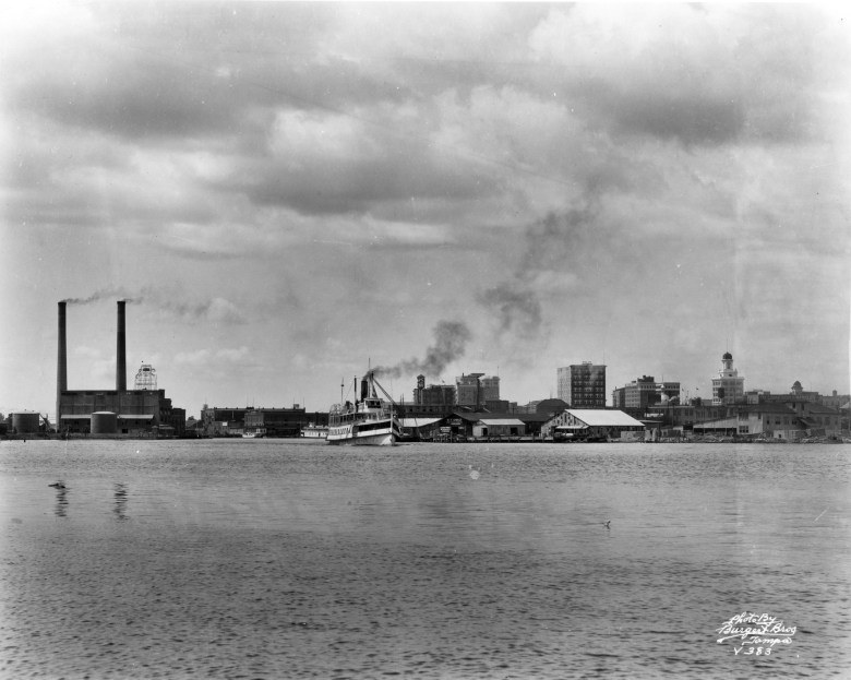 A waterfront with industrial buildings and smokestacks in the background, taken from across the water. Several boats are docked along the shore under a cloudy sky.