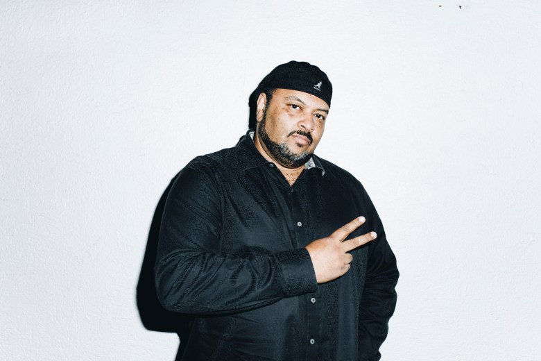 A portrait photograph by Gary Go showing musician and producer Egyptian Lover posing indoors at the Fauna Primavera Festival at Espacio Broadway in Santiago, Chile, on November 10, 2018. Captured in a medium close-up shot centered against a clean white backdrop, Egyptian Lover gestures a peace sign with his left hand while looking toward the camera. He wears a dark, long-sleeved button-down shirt paired with a dark Kangol beret cap