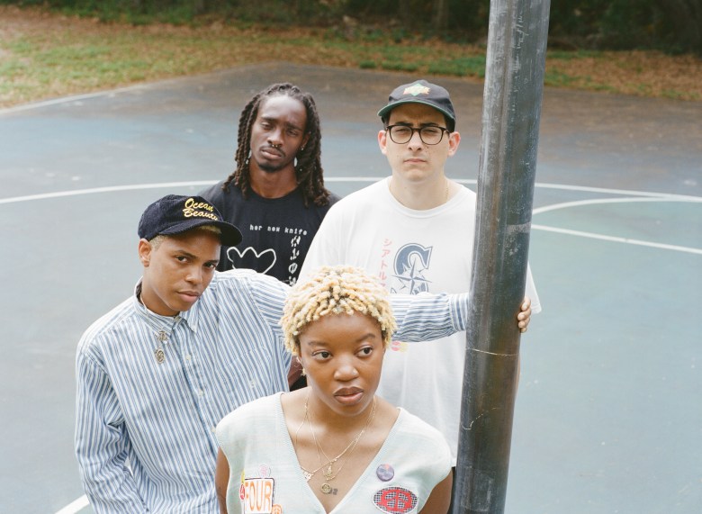 A high-angle outdoor group portrait by Zuri Moult showing four members of Fesssi K. standing together on an outdoor basketball court. Arranged from left to right, Vonne Parks stands on the far left wearing a dark corduroy cap embroidered with "Ocean Beauty" and a light blue striped button-down shirt; Micah Jno Baptise stands in the back left wearing a black t-shirt printed with "her new knife"; Noel Colon stands in the back right wearing glasses, a black baseball cap, and a white graphic t-shirt while resting his hand on a metallic pole; and Zoe Robinson stands in the center foreground wearing a light grey sweater vest adorned with patches and layered gold necklaces, featuring short blonde styled hair.