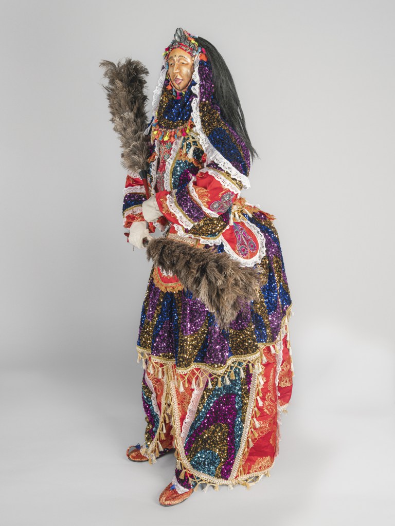 A full-length studio photograph of the Jollay Society 'Fairy' Masquerade Ensemble created in 2023 by Sheku 'Goldenfinger' Fofanah and tailor Dezo Sesay, commissioned for the Fitchburg Art Museum. Displayed against a plain light grey backdrop, the life-size masquerade figure stands facing left in a richly detailed, multi-colored garment decorated with metallic sequins, red lace trims, and gold fringe. The figure wears a carved wooden face mask under a dark veil and headpiece, holding a large grey feathered flywhisk broom across its torso while standing in red embroidered shoes.