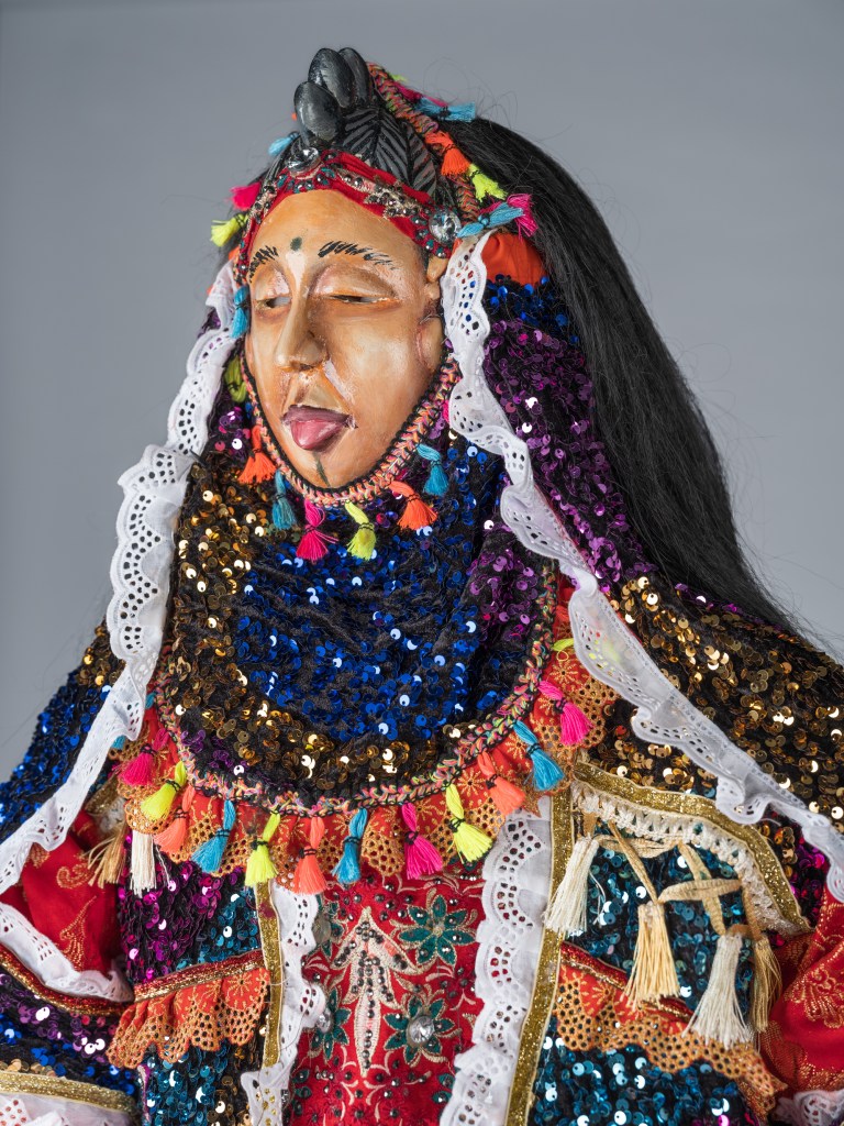 A detailed close-up portrait photograph of the Jollay Society 'Fairy' Masquerade Ensemble created in 2023 by Sheku 'Goldenfinger' Fofanah and tailor Dezo Sesay, commissioned for the Fitchburg Art Museum. Set against a plain light grey backdrop, the tight shot focuses on the carved wooden face mask featuring a slight smile, extended tongue, and dark carved hair, framed by white lace borders and colorful tassels in pink, yellow, and blue. The intricate, layered garments are adorned with blue, gold, and purple metallic sequins, embroidered red lace, gold trim, and hanging white tassels.