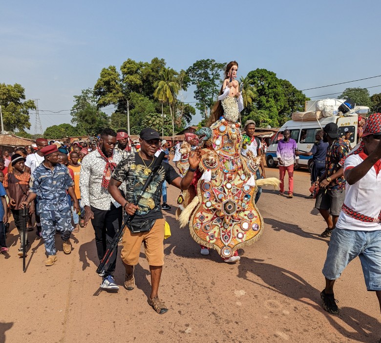 An outdoor photograph showing Sheku 'Goldenfinger' Fofanah leading a procession with his Mami Wata devil during the Massaboni Ordehlay procession in Lunsar, Sierra Leone, on December 26, 2022. Captured in a full-length wide shot along a dirt road under a clear blue sky, Fofanah walks forward wearing a short-sleeved camo shirt, brown shorts, sandals, a black baseball cap, and a crossbody bag while holding a wooden staff. Immediately behind him, a towering Mami Wata masquerader wears an elaborate, ornate suit embellished with circular mirror discs, gold fringe, and multi-colored patterned textiles, topped with a mannequin-like headpiece wearing a long dark wig. Onlookers and procession participants surround them along the dusty road framed by lush green trees in the background.