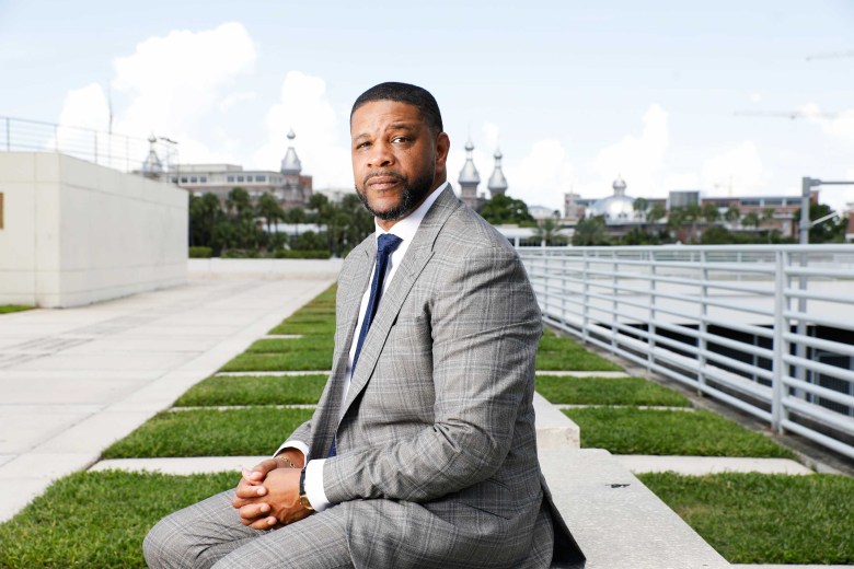A photograph showing political candidate Gary Hartfield sitting outdoors on a concrete bench at Kiley Garden in Tampa, Florida. Captured in a medium three-quarter shot, Hartfield sits with his hands clasped together in his lap, looking directly forward toward the camera. He wears a grey plaid suit with a white dress shirt and dark blue tie. Behind him, the open-air plaza features green lawn strips, a white concrete wall, metal railings, and the distant rooftops and spires of downtown Tampa under a partly cloudy sky.