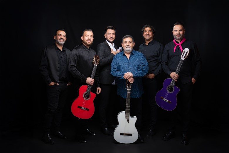 A studio promotional photograph of the music group Gipsy Kings posing against a plain black background. Six musicians stand in a row dressed in dark collared shirts and trousers under studio lighting. In the center, lead guitarist Tonino Baliardo wears a bright blue button-down shirt and holds a white acoustic guitar pointing downward. Flanking him on either side, band members pose holding vibrant colored acoustic guitars—a red guitar on the left and a purple guitar on the right—with one member on the far right wearing a bright red neck scarf.