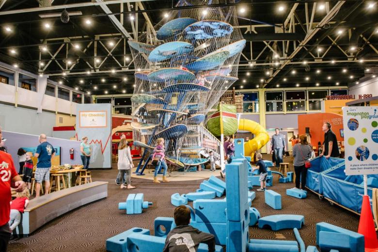 A wide interior photograph showing young children and families playing inside the main exhibit hall at Great Explorations Children's Museum in St. Petersburg, Florida. In the center, a multi-story climbing structure wrapped in netting features stacked blue platforms extending toward a high ceiling with exposed metal trusses and track lighting. In the foreground, large blue foam building blocks scattered across a carpeted floor allow children to build structures, alongside a yellow slide and interactive play stations illuminated by bright overhead lights.