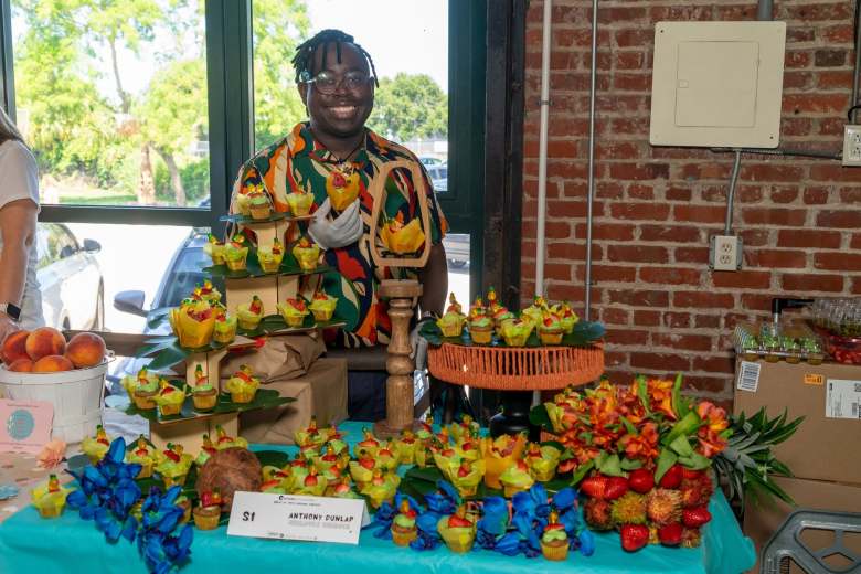 A photograph showing baker Anthony Dunlap standing behind an elaborate cupcake display table at the Great St. Pete Cupcake Contest inside the Morean Center for Clay in St. Petersburg, Florida, on August 24, 2024. Captured in a medium shot, the smiling baker wears a colorful patterned tunic, glasses, and a face mask around his chin while standing behind a teal tablecloth filled with dozens of decorated cupcakes arranged on multi-tiered wooden and orange cake stands, flanked by vibrant floral arrangements and set against an exposed red brick wall.