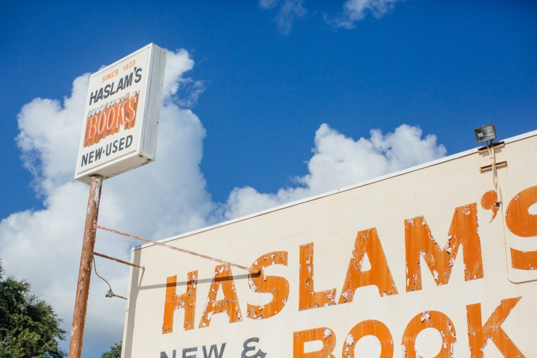 A low-angle exterior photograph showing the signage for Haslam's Book Store in St. Petersburg, Florida, captured on July 8, 2016. Set against a bright blue sky with fluffy white clouds, the primary shot features orange weathered lettering on the building's facade reading "HASLAM'S NEW & USED BOOKS." To the left, a tall wooden pole holds a raised rectangular white sign reading "HASLAM'S BOOKS NEW & USED" with orange lettering.