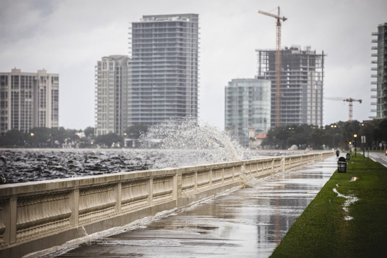 A wide exterior photograph showing storm surge crashing over the seawall along Bayshore Boulevard in Tampa, Florida, during Hurricane Helene on September 26, 2024. Captured at a wide angle along the wet, paved pedestrian walkway, a large wave sprays white foam over the concrete balustrade onto the sidewalk. In the background across dark, choppy waters, high-rise residential buildings and construction cranes stand under a grey, overcast storm sky.