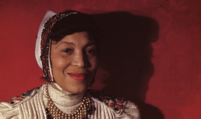 A color Kodachrome portrait photograph taken by Carl Van Vechten showing author Zora Neale Hurston on July 18, 1946. Captured in a close-up portrait against a rich red backdrop, Hurston smiles warmly toward the right of the frame, casting a strong shadow behind her. She wears an off-white cream garment adorned with beaded trim at the collar and shoulders, multi-strand beaded necklaces, and a light-colored bonnet tied beneath her chin with an embellished, jewel-toned netted overlay across her dark hair.