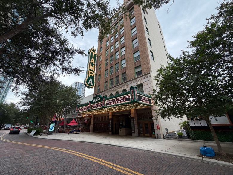 A wide exterior photograph showing the historic Tampa Theatre building on a brick-paved street in downtown Tampa, Florida, on August 20, 2026. Captured from across the street at a low angle, the mid-rise building features a tall vertical neon sign spelling "TAMPA" mounted along its side and an illuminated marquee marquee above the entrance overhang displaying featured movie titles. Green trees flank the street on both sides, with brick pavers curving through the foreground under an overcast sky.