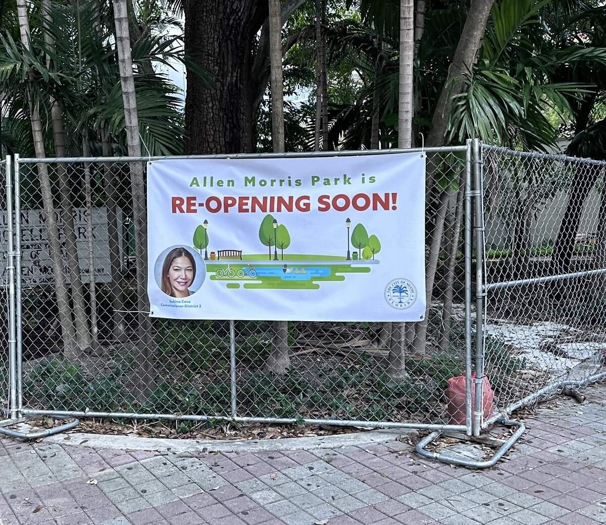 A banner showing then-Miami Commissioner Sabina Covo’s photo was installed outside Allen Morris Brickell Park in 2023 announcing its reopening after years of litigation with the Allen Morris Company. The reopening was temporary and the park shut down to the public again in July 2026. The company plans to open a restaurant at the site and the land is zoned for a high-rise.