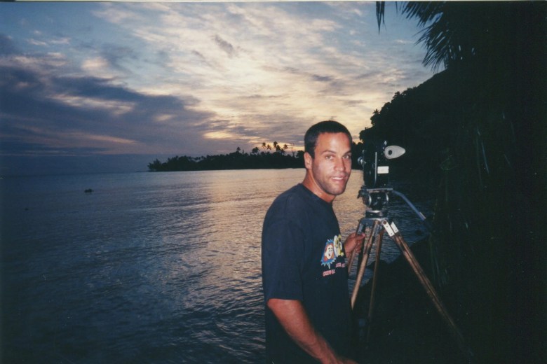 A photograph taken by K. Johnson showing musician and filmmaker Jack Johnson on location filming the surf documentary 'Thicker Than Water' in Tahiti circa June 1998. Captured at dusk in a three-quarter shot along the shoreline, Johnson stands smiling toward the camera in a dark short-sleeved t-shirt next to a film camera mounted on a tripod. Behind him, calm ocean waters reflect a colorful sunset sky filled with soft clouds, framed by dark palm trees along the coast on the right.