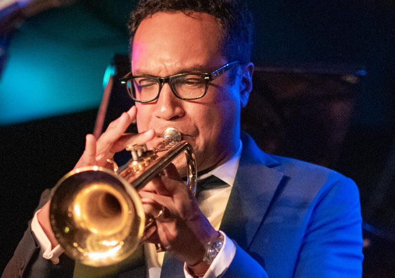 A performance photograph by Stephen Splane showing jazz trumpeter James Suggs performing live at the Sarasota Jazz Festival inside the Hyatt Regency Sarasota in Sarasota, Florida, on March 6, 2019. Captured in a close-up shot under dramatic blue stage lighting, Suggs plays a brass trumpet angled toward the left, wearing a blue suit jacket, a white collared shirt with a dark tie, dark-framed eyeglasses, and a wristwatch on his left wrist.