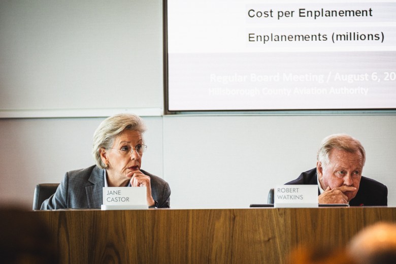 A photograph by Dave Decker showing Tampa Mayor Jane Castor and board member Robert I. Watkins seated at a long wooden dais during a meeting at Tampa International Airport on August 6, 2026. On the left, Castor wears a grey suit jacket over a light collared shirt and rests her chin on her hand beside a printed nameplate reading "JANE CASTOR." On the right, Watkins wears a dark suit jacket with his hands clasped under his chin beside a nameplate reading "ROBERT WATKINS." Behind them, a presentation screen displays text reading "Cost per Enplanement / Enplanements (millions)."