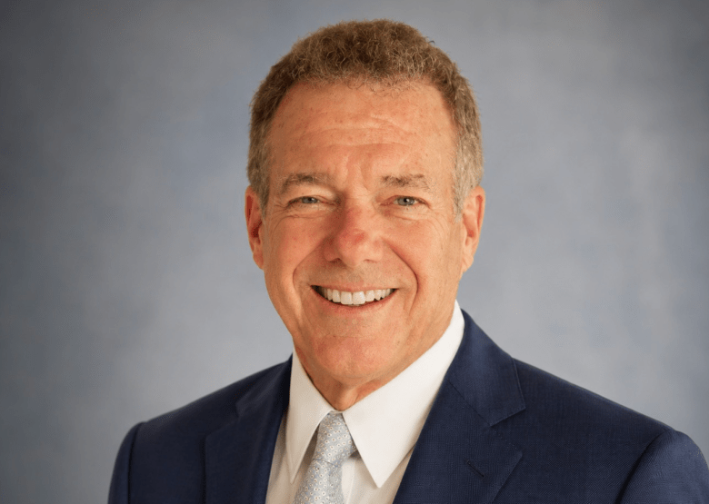 A formal studio headshot photograph of Jeff Patterson posing for his Tampa City Council campaign. Positioned center frame against a mottled grey studio backdrop, Patterson smiles brightly directly into the camera lens in a close-up portrait. He wears a dark navy blue suit jacket paired with a crisp white collared dress shirt and a light silver patterned necktie, illuminated under soft overhead studio lighting.