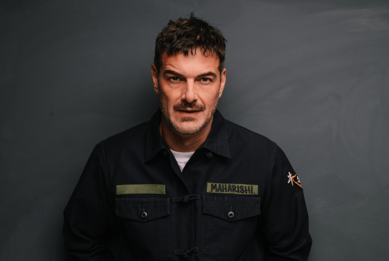 A promotional portrait of electronic music producer and DJ John Graham, aka Quivver. Captured in a medium close-up shot against a dark grey background, Graham looks directly into the camera with a subtle expression. He wears a dark jacket featuring a white undershirt underneath and a woven yellow chest patch reading "MAHARISHI" over his left pocket.