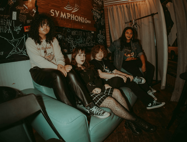A promotional group photograph of Tampa band Just Courtesy posing together in a dimly lit indoor space with chalk-covered walls and a wooden sign reading "SYMPHONIC." Captured in a wide shot, the four band members sit together along a teal leather couch and adjacent seating. From left to right: a member with dark curly hair wears a long-sleeve graphic shirt and dark leather pants; a second member sits in a dark top, plaid skirt, patterned tights, and high-top sneakers; a third member with wavy red hair wears a dark graphic t-shirt; and a fourth member in a dark denim jacket sits on the armrest wearing black sneakers with white laces under warm, moody interior lighting.
