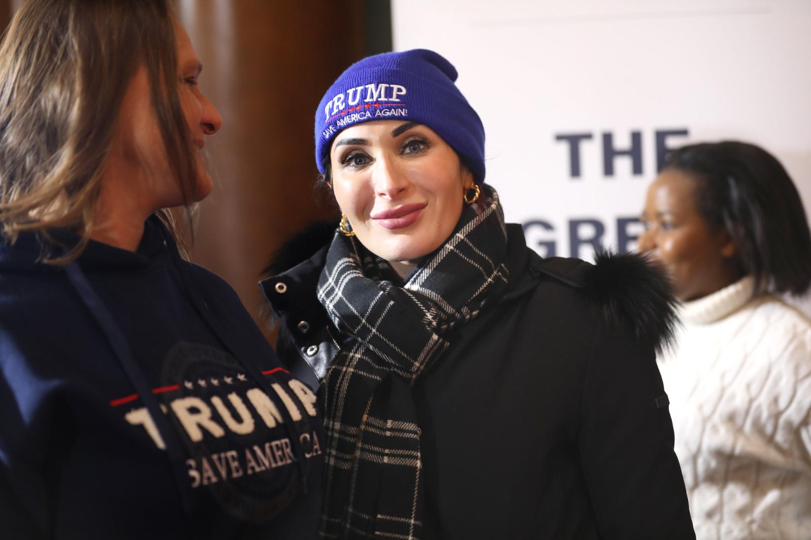 Laura Loomer wears a blue beanie looking at the camera with a closed-mouth smile. She is wearing a black scarf and a puffer coat with a fur hood.