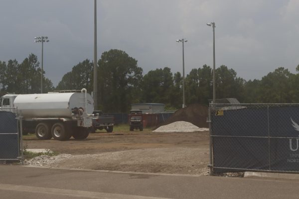 An outdoor construction area at a university, seen behind a chain-link fence. On the left, a large white water truck is parked. In the background, there is a golf cart, a pickup truck, a pile of white gravel next to a dirt mound, and a small utility building. Tall light poles and dense green trees stand under an overcast sky.