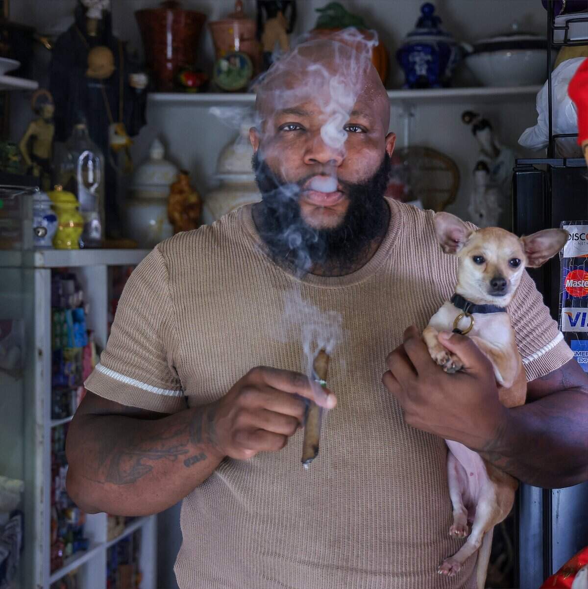 Universal Santeria priest Hector Giovanni Pizarro, known as “Oba Ti Lemi” to practitioners, holds his dog while preparing to bless his botanica with cigar smoke as he faces rising rent and the aftermath of a water-damaged storefront in Fort Lauderdale, Monday, July 20, 2026.