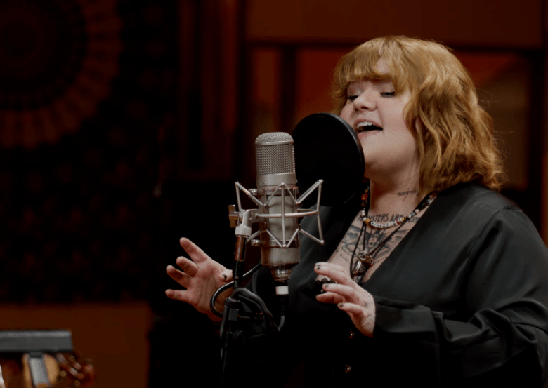 A screenshot from singer-songwriter MaKenzie's YouTube video "Velvet (Live at EastWest Studios)." Captured in a medium close-up shot inside a dark recording studio, MaKenzie sings passionately with her eyes closed into a silver studio condenser microphone fitted with a black pop filter. She wears a black long-sleeved shirt over a patterned top, stacked layered necklaces, and short wavy auburn hair with bangs, softly lit by ambient studio lighting.