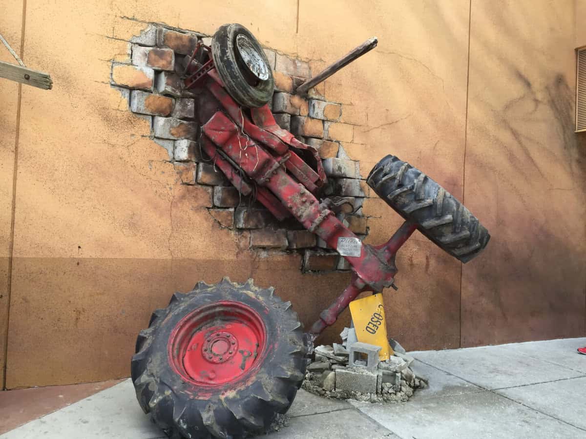 A red tractor bursts dramatically through a brick wall, with scattered debris and shattered bricks engulfing its wheels—reminiscent of the heart-pounding spectacle you’d experience on Universal Orlando’s iconic Twister Ride.