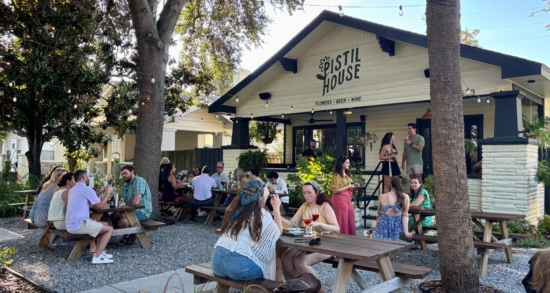 An outdoor seating area at Pistil House, showing groups of people seated at wooden picnic tables under string lights and trees while “Porch Tunes” are performed in front of the black-and-white building.