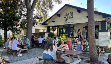 An outdoor seating area at Pistil House, showing groups of people seated at wooden picnic tables under string lights and trees while “Porch Tunes” are performed in front of the black-and-white building.