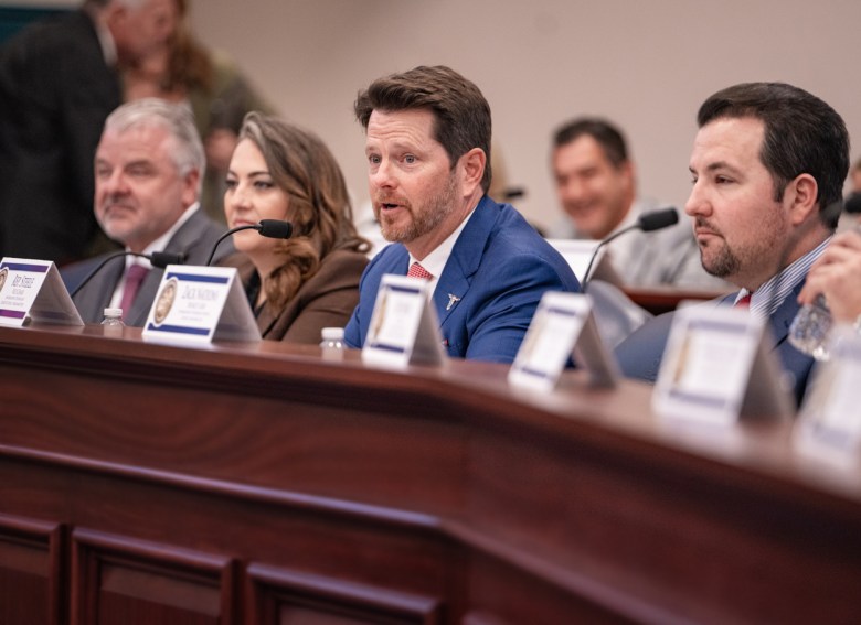 Representative Kevin M. Steele speaks at a legislative committee hearing, wearing a blue suit and red tie, seated at a long, dark wood desk with other members;