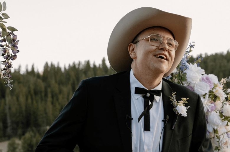 A photograph of comedian and author Sam Tallent posing outdoors in western-style formal wedding attire. Positioned center frame in a medium close-up shot, Tallent looks upward and to the right with a wide, joyful smile. He wears a light beige cowboy hat, thin gold-framed eyeglasses, a black suit jacket, a white collared dress shirt, a floral boutonnière pinned to his lapel, and a black string bolo tie. In the background, soft outdoor daylight illuminates a lush green pine tree forest beneath floral wedding arrangements of white and lavender flowers.