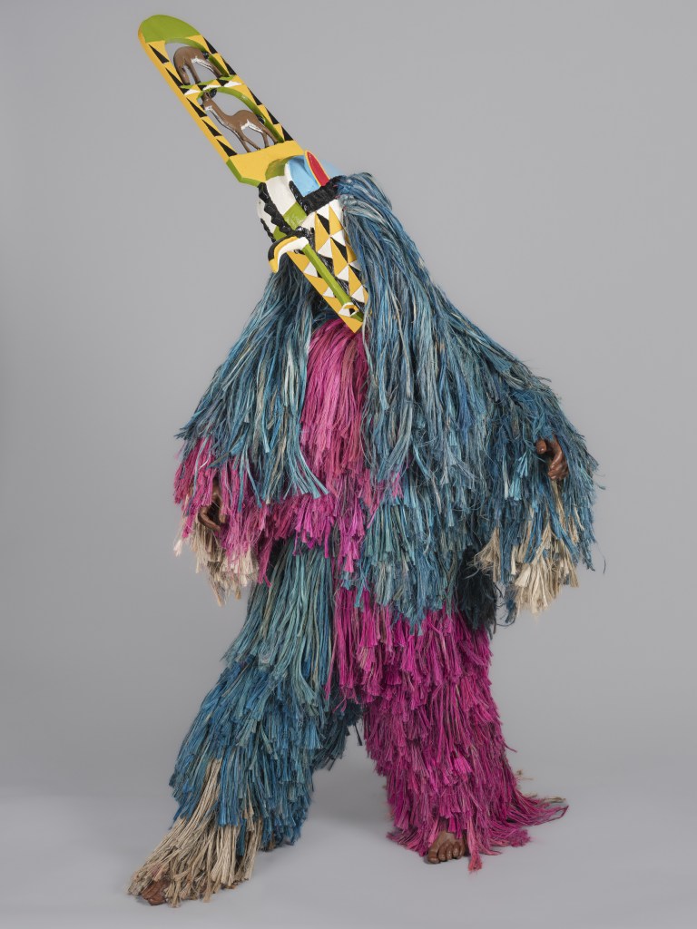A studio photograph of the full life-size Kimi Masquerade Ensemble artwork created in 2022 by David Sanou (headpiece carved in the studio of André Sanou, with an anonymous body maker), commissioned for the Fitchburg Art Museum. Displayed against a plain neutral grey backdrop, the figure stands in a full raffia-fiber suit featuring alternating panels of vibrant magenta, slate blue, and natural tan fibers. Attached to the head is a geometric, carved wooden mask topped with an angled, multi-colored plank extension painted in black, white, yellow, red, and green geometric patterns with bird-like motifs.
