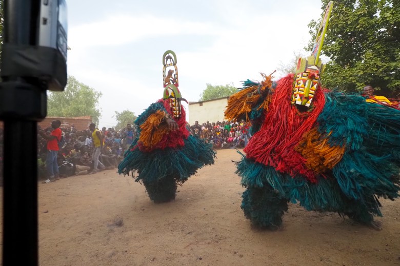 An outdoor photograph showing a pair of Kimi masks with headpieces carved by David Sanou in the studio of André Sanou performing greetings in the Bindougosso district of Bobo-Dioulasso, Burkina Faso, on May 3, 2022. Captured in a dynamic low-angle wide shot in a dusty open-air village square, two masqueraders dance in voluminous suits constructed from bright red, teal, and gold raffia fibers, topped with carved geometric wooden headpieces featuring painted patterns and tall vertical crest extensions. In the background, lead griot Tchiedo plays a traditional drum alongside a crowd of seated spectators watching the performance under an open sky.