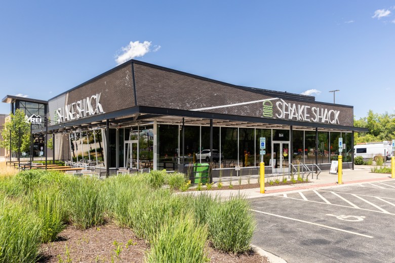 A photograph by Joseph Hendrickson showing the exterior of a modern Shake Shack restaurant location in Vernon Hills, Illinois, on June 26, 2024. Captured from a wide exterior angle, the single-story dark brick and glass building features large white "SHAKE SHACK" signage with green burger logos mounted on both its front and side facades. In the foreground, green ornamental grasses, native plants, and manicured landscaping frame the building, while an asphalt parking lot with accessible spaces and yellow safety bollards sits under a clear blue sky in bright daylight.
