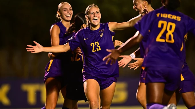 Female soccer players in purple uniforms celebrate together on a night game field, arms raised in joy.