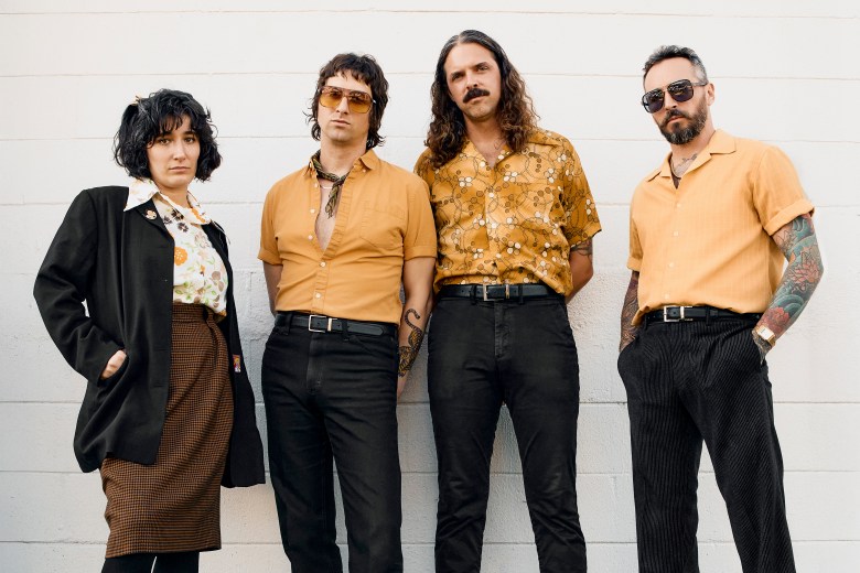A full-length promo photograph by Joey Clay showing four members of the band Soft Cuff standing side-by-side against a white brick wall. From left to right: Christina Sanchez stands with one hand in her coat pocket wearing a black blazer over a floral collared shirt, dark patterned skirt, and tights; Michael Bostinto stands in a mustard-yellow button-down shirt with a dark neck scarf, dark trousers, and tinted sunglasses; Brian Schanck stands with long shoulder-length hair and a mustache wearing a gold patterned short-sleeve button-down shirt tucked into dark pants; and Shane Shuch stands on the far right wearing sunglasses, a peach short-sleeve collared shirt, dark pants, and displaying sleeve tattoos on his arms under bright, even daylight.