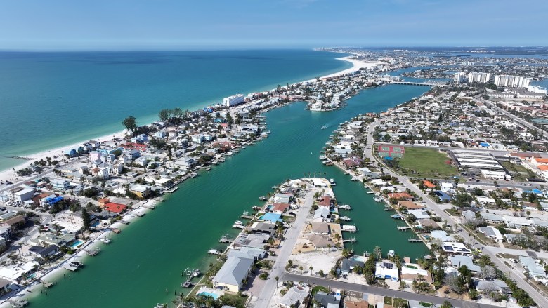 A high-altitude aerial drone photograph by ByDroneVideos showing a panoramic view of St. Pete Beach, Florida. The shot looks northwest along the barrier island, highlighting a vibrant turquoise intracoastal waterway flowing between residential neighborhoods of coastal homes, boat docks, and palm trees. To the left, white sand beaches meet the clear blue waters of the Gulf of Mexico under a bright, cloudless blue sky extending to the horizon in natural daylight.