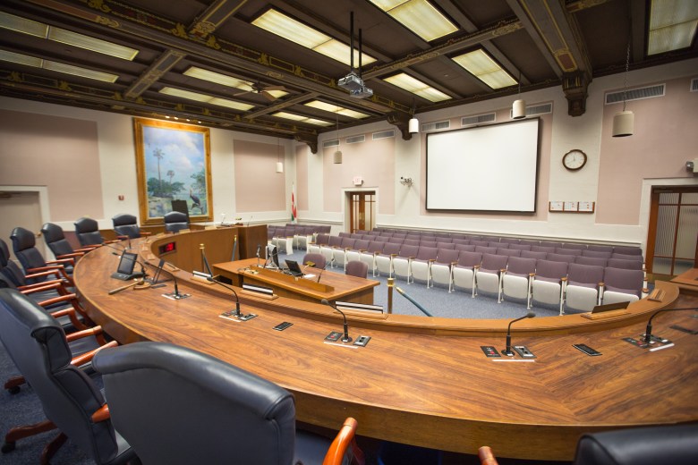 A wide-angle interior photograph showing the empty St. Petersburg City Council Chambers in St. Petersburg, Florida, on November 18, 2014. Captured from behind a curved, polished wooden dais lined with dark leather executive chairs, microphones, and computer monitors, the room features carpeted flooring and rows of grey spectator chairs facing a central presentation screen on the back wall. Coffer-style wooden ceiling panels with warm lighting fixtures overhead and a framed tropical landscape painting on the left wall illuminate the municipal assembly room.