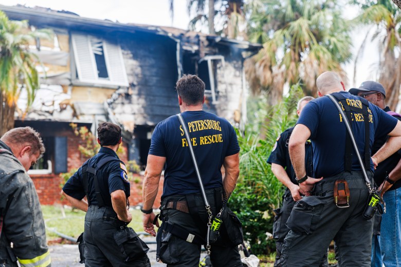 A photograph of St. Petersburg Fire Rescue personnel assessing storm damage and flood recovery efforts in the Shore Acres neighborhood of St. Petersburg, Florida, on August 31, 2023, following Hurricane Idalia. Captured in a medium outdoor shot, two firefighters wearing navy St. Petersburg Fire Rescue t-shirts and dark trousers stand alongside a local resident outside a residential house. Debris, discarded flood-damaged items, and water residue line the lawn and concrete driveway under daylight