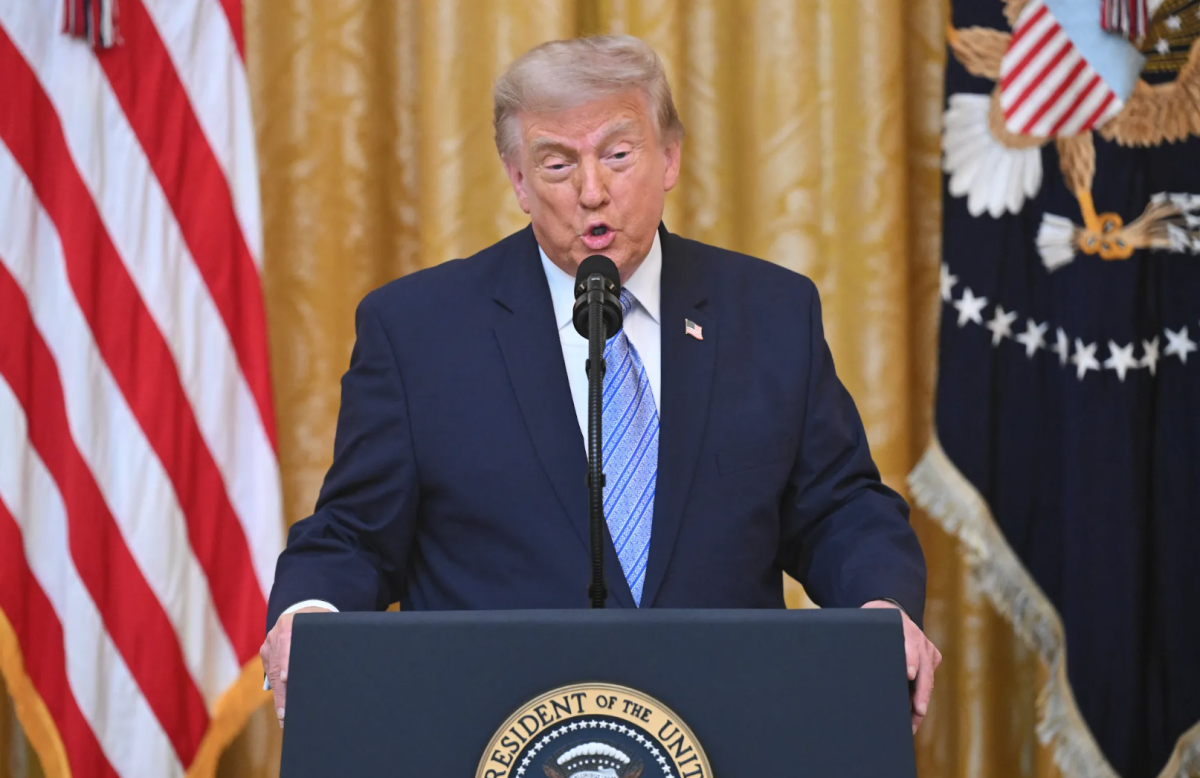 Donald Trump speaking at a podium bearing the seal of the President of the United States, flanked by the American flag and the Presidential flag.